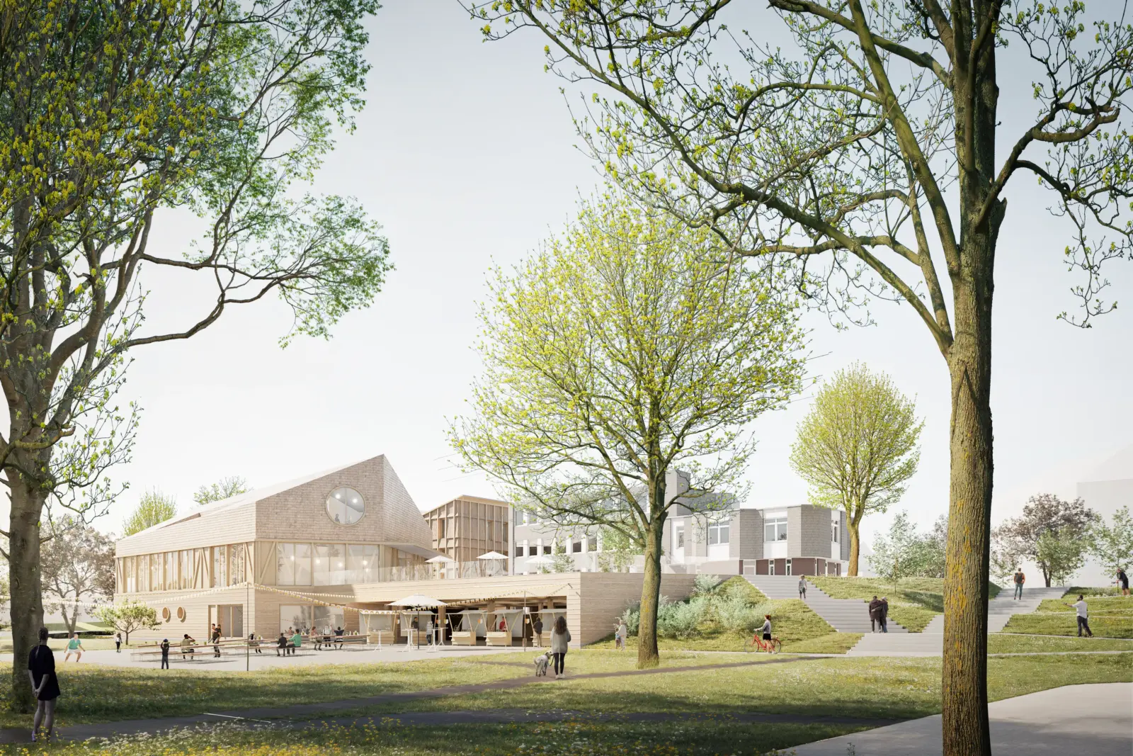 Das Gebäude zeigt eine Holzfassade mit rundem Fensterelement und einem markanten Giebeldach, im Hintergrund schließen sich weitere Baukörper mit heller Putzfassade an.