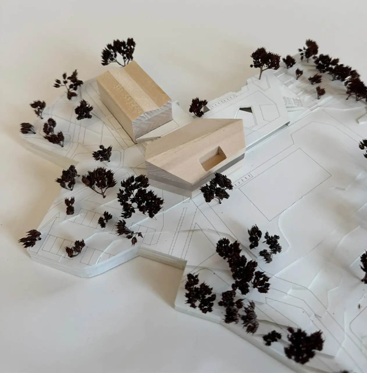 Modellfoto mit zwei holzfarbenen Baukörpern auf einem weißen Lageplanmodell mit Straßenführung und Bäumen.