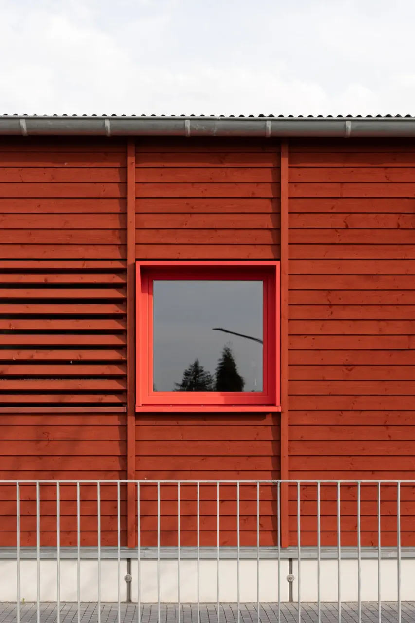 Fassadendetail mit horizontaler Holzverschalung und quadratischem Fenster in rotem Rahmen.