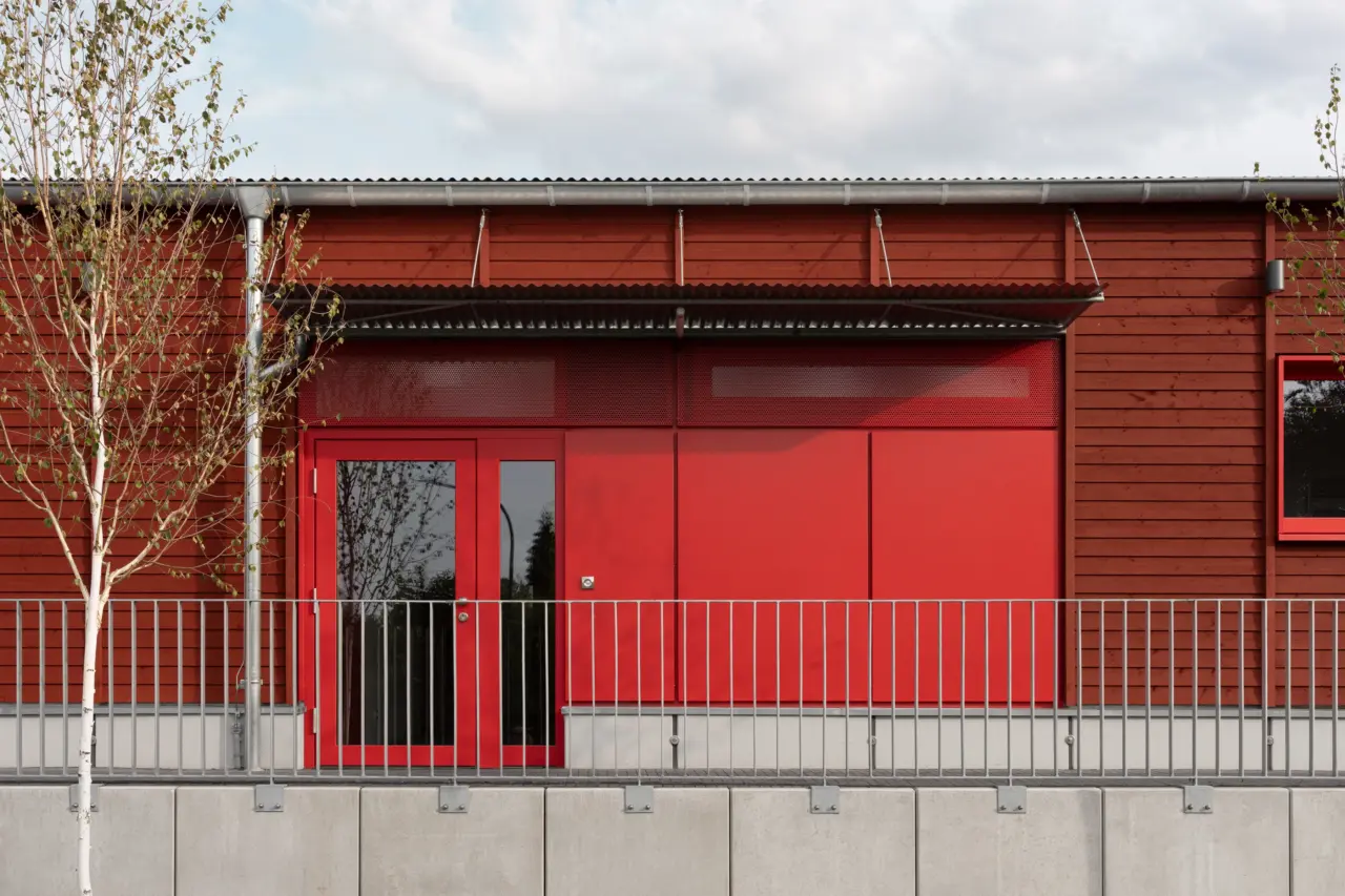 Rot verkleidete Fassadenöffnung mit Glastür an einem Gebäude mit horizontaler Holzverschalung.
