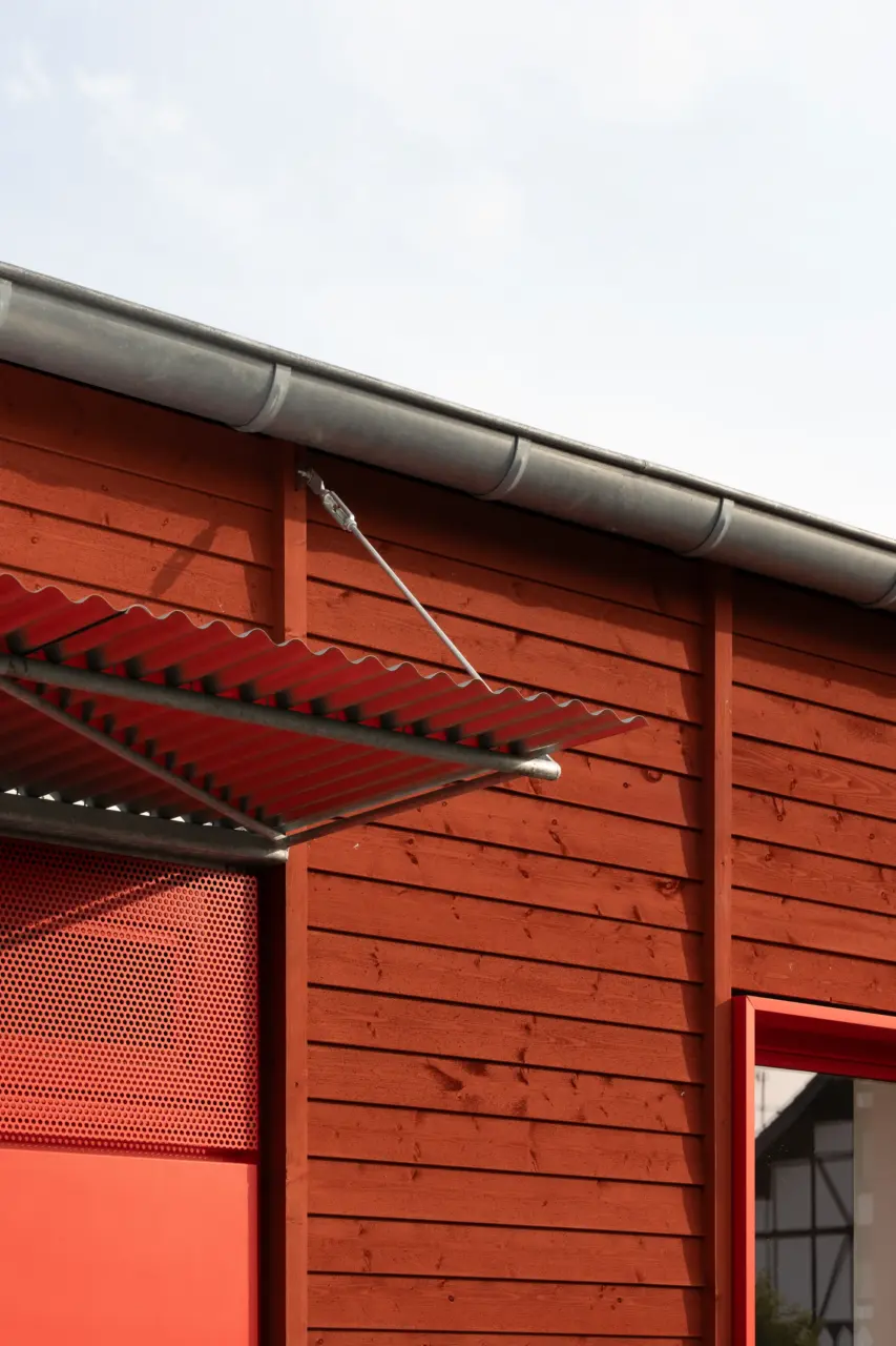 Detailansicht der Holzfassade mit rotem Wellblechvordach und verzinkter Dachtraufe.