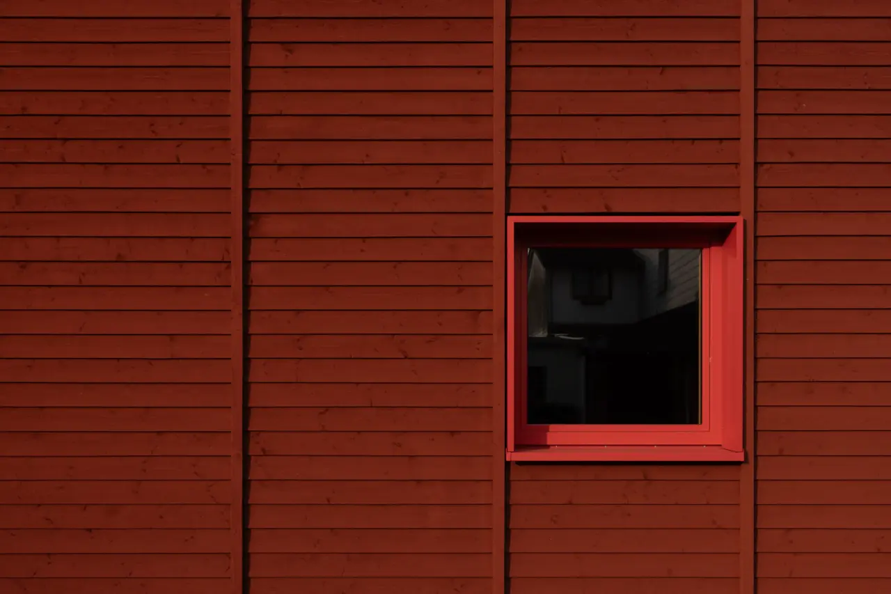 Fassadenausschnitt mit horizontaler Holzschalung in Rotbraun und quadratischem Fenster mit rotem Rahmen.