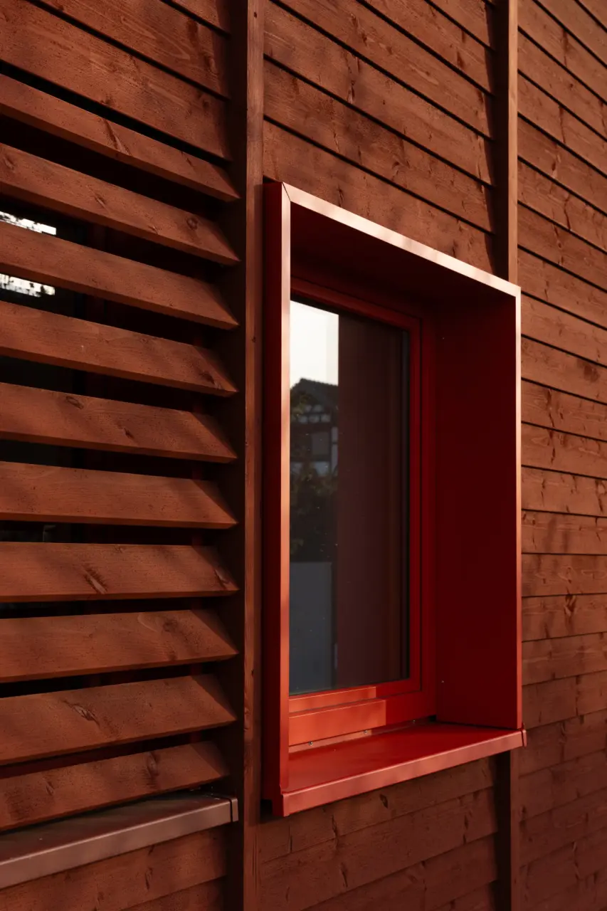 Fensterausschnitt in einer horizontalen Holzlattenfassade mit rot eingefasster Laibung und Fensterbank aus Metall.