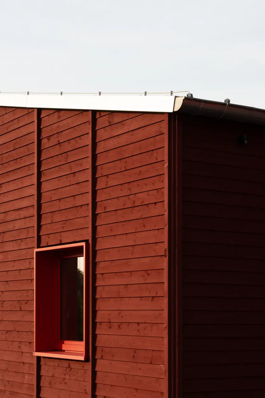 Gebäudeecke mit rot lasierter Holzfassade aus Rhombusleisten und einem tief eingeschnittenen Fenster mit rotem Rahmen.