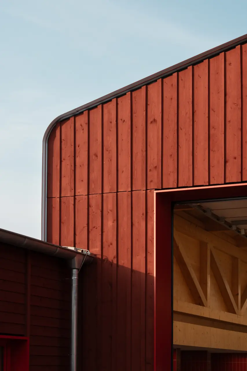 Fassadendetail mit vertikaler Holzschalung und abgerundeter Ecke, im Hintergrund eine geöffnete Toröffnung mit Holzkonstruktion.