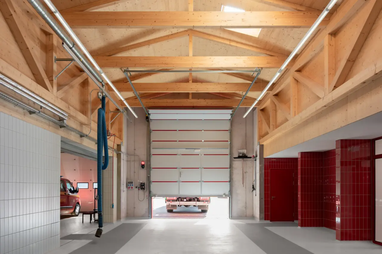 Innenraum einer Fahrzeughalle mit sichtbarem Dachtragwerk aus Holz und geöffnetem Sektionaltor, dahinter ein Fahrzeug.