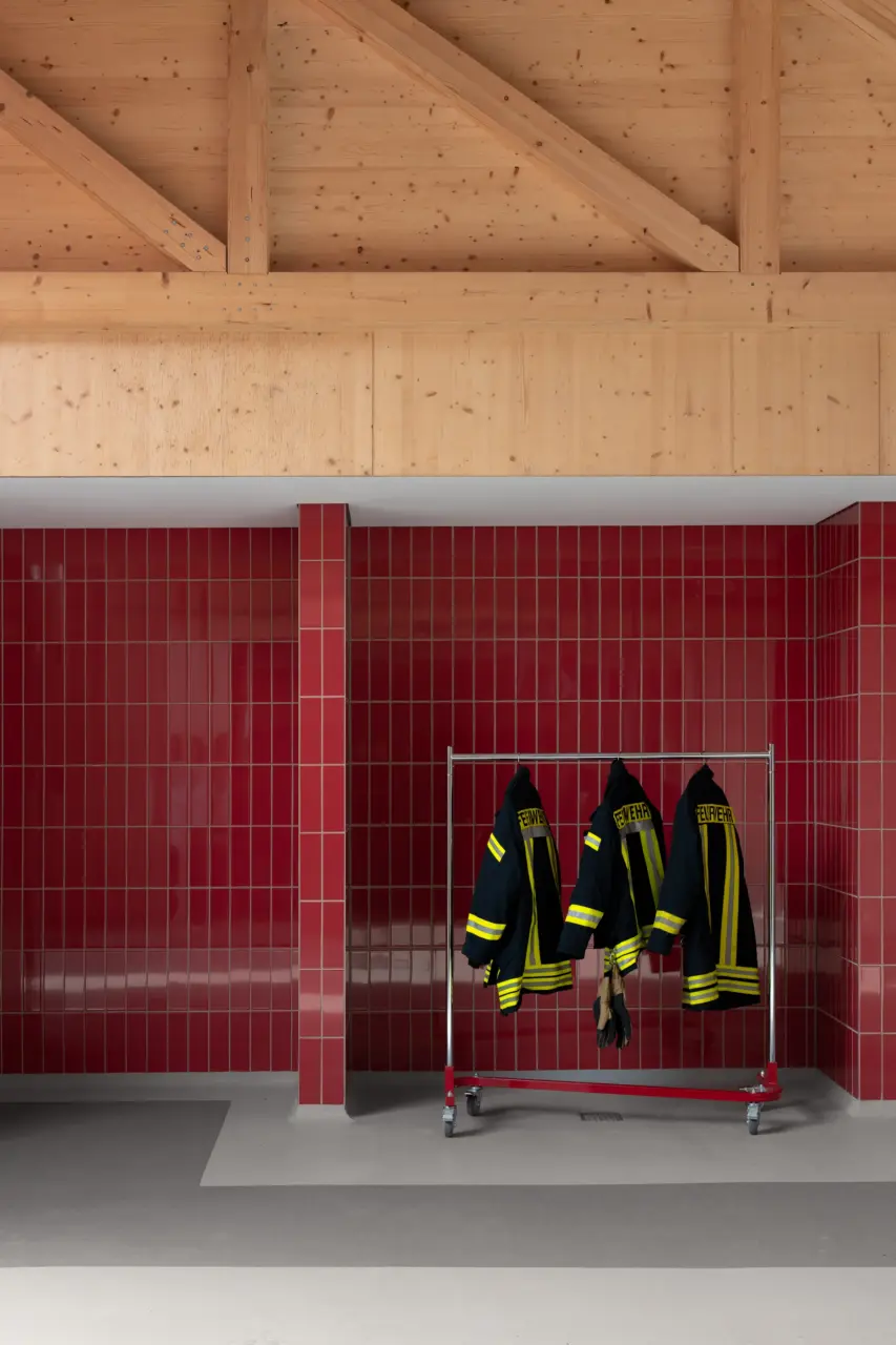 Rollbares Gestell mit drei Feuerwehrjacken vor rot gefliester Wand unter offenem Holzdachstuhl.