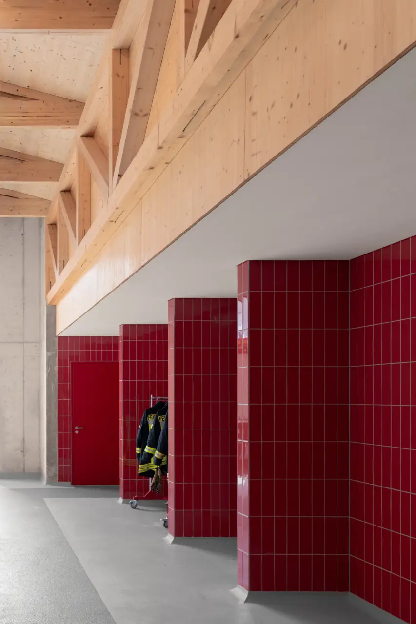 Innenraum einer Feuerwache mit rot gefliesten Umkleidenischen unter einem sichtbaren Holzdachtragwerk.