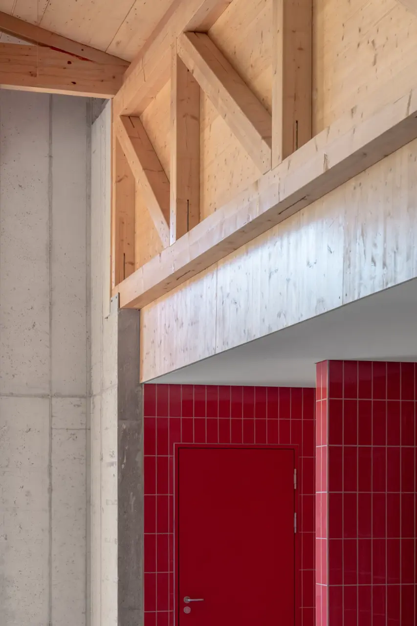 Im Innenraum zeigt sich ein Dreiecksfachwerk aus hellem Holz unter der Dachschräge, links begrenzt von einer Sichtbetonwand. Darunter schliesst eine Wand mit roten Fliesen an, in die eine ebenfalls rote Tür eingelassen ist.