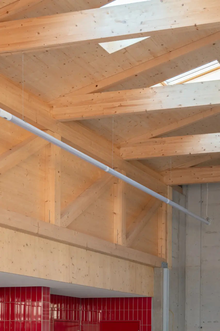 Innenraum mit sichtbarer Holzkonstruktion aus Dachbalken, Dreiecksfenstern im Dach und rot gefliester Wand darunter.