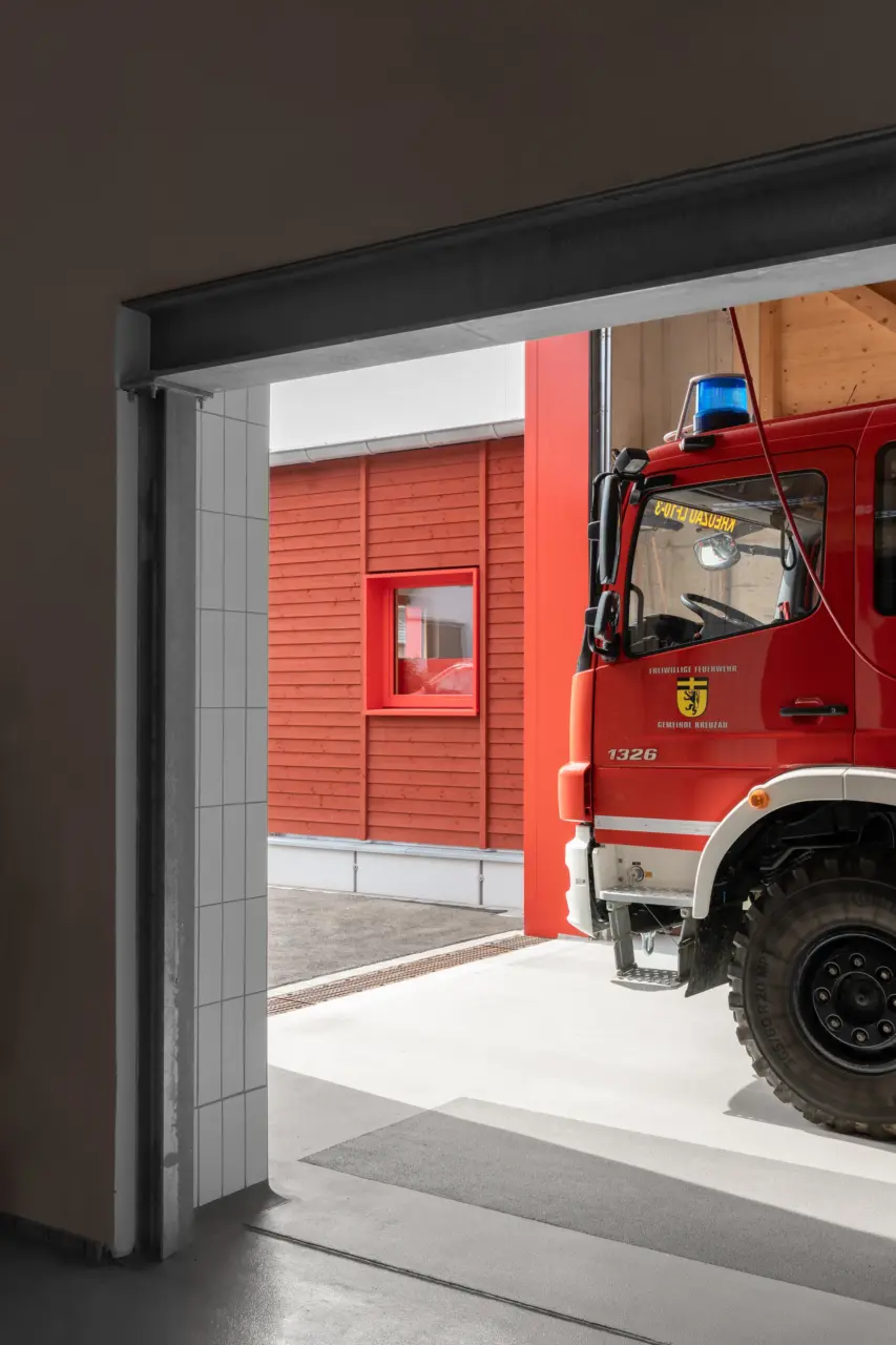 Blick durch ein Rolltor auf ein Feuerwehrfahrzeug, im Hintergrund eine Fassade aus roter Holzschalung.