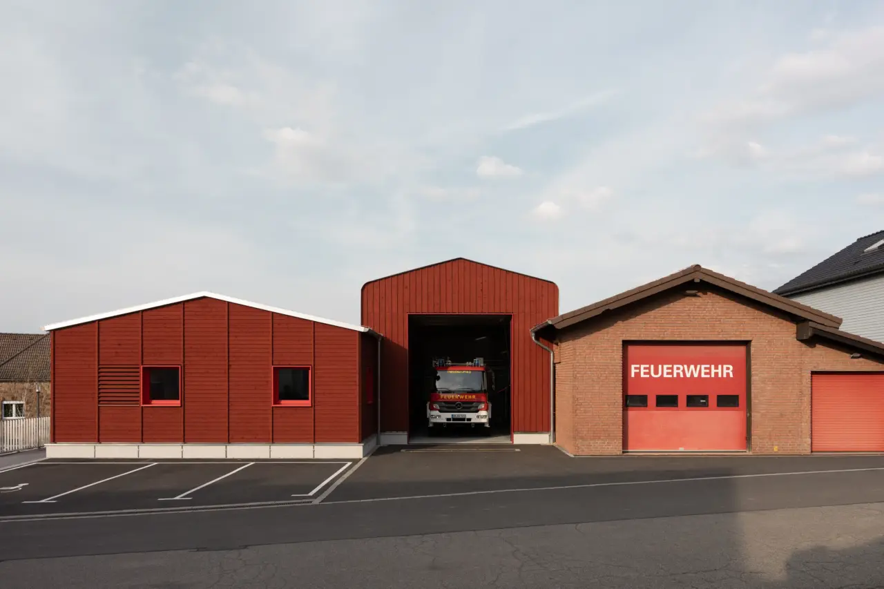 Feuerwache mit rotbrauner Holzfassade und geöffnetem Tor, in dem ein Feuerwehrfahrzeug steht, daneben ein Backsteinbau