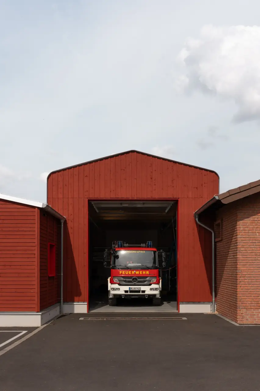 Außenansicht einer Feuerwache mit rot verkleideter Holzfassade und geöffnetem Tor, in dem ein Feuerwehrfahrzeug steht.