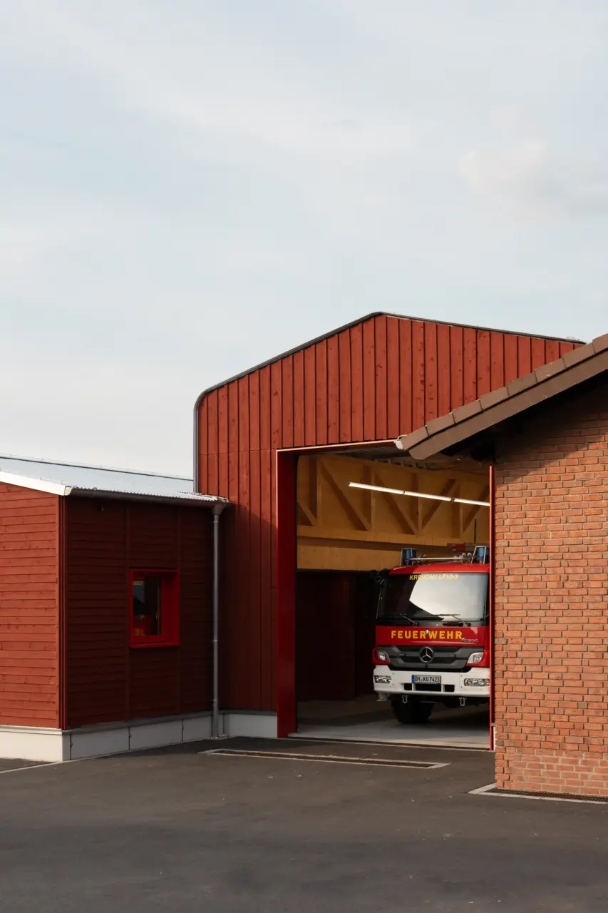 Geöffnetes Rolltor einer Feuerwache mit rotbrauner Holzfassade gibt Blick auf ein Löschfahrzeug frei.