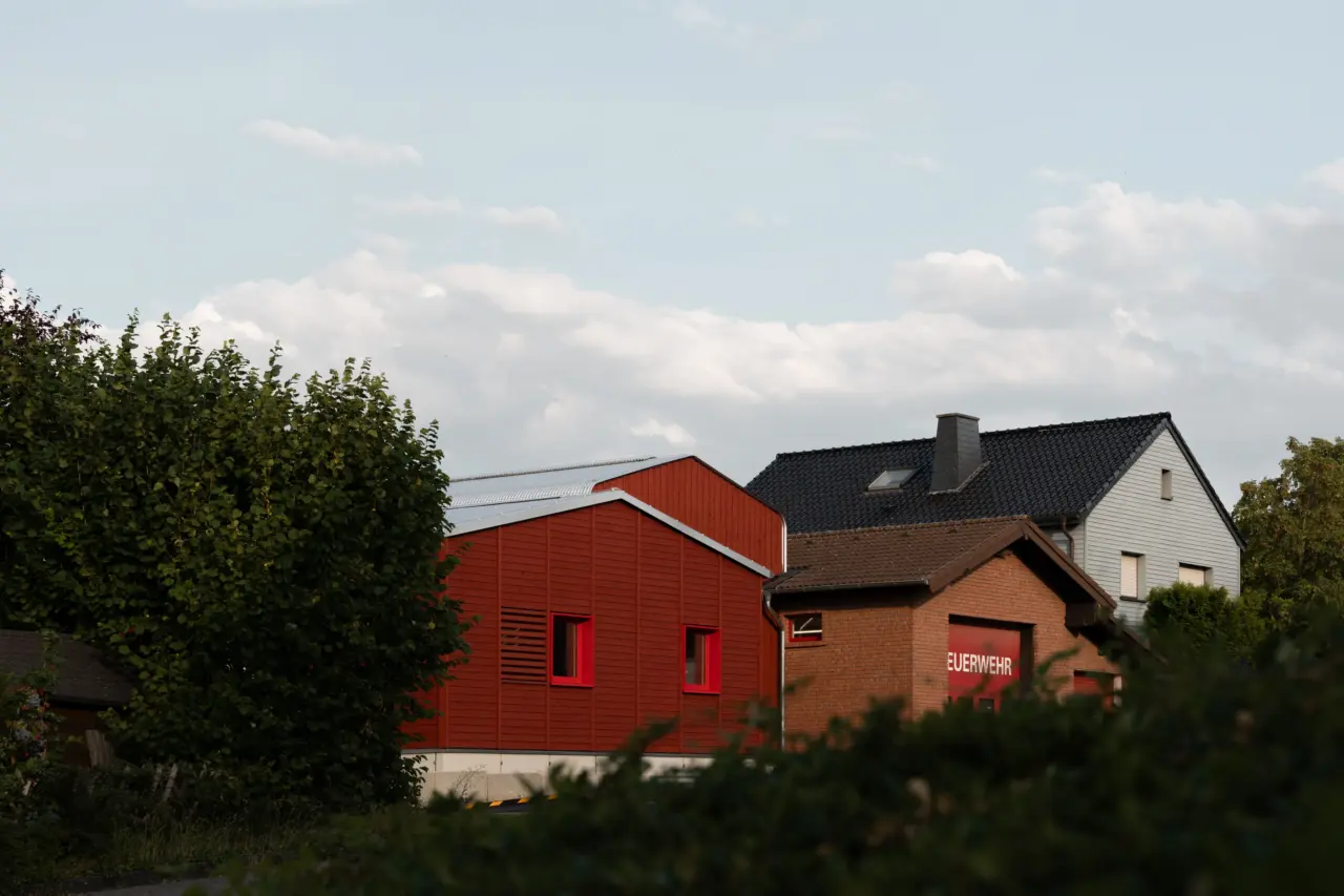Feuerwache mit rot verkleidetem Neubau und angrenzendem Ziegelbau, hinter Bäumen und Sträuchern hervorragend.