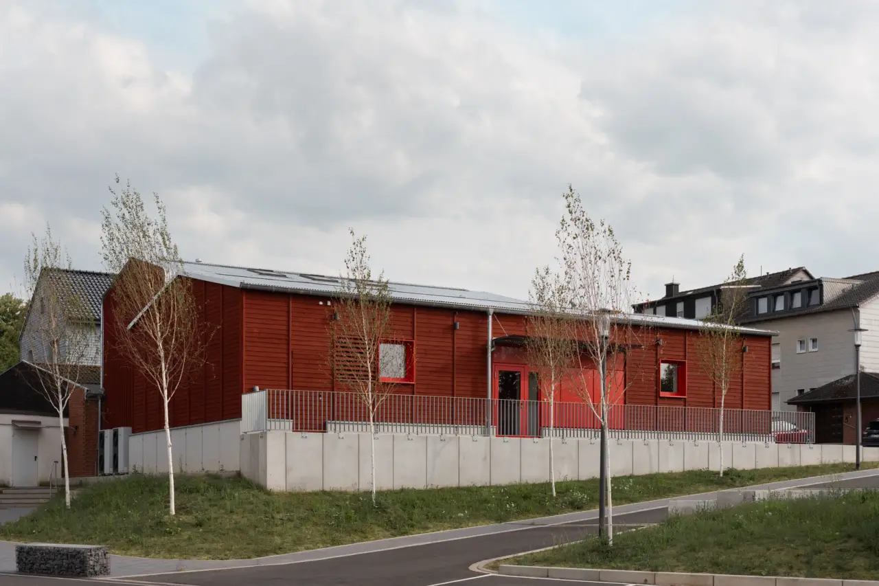 Außenperspektive der Feuerwache mit rot lasierter Holzfassade und Sockel aus Sichtbeton, davor junge Birken.
