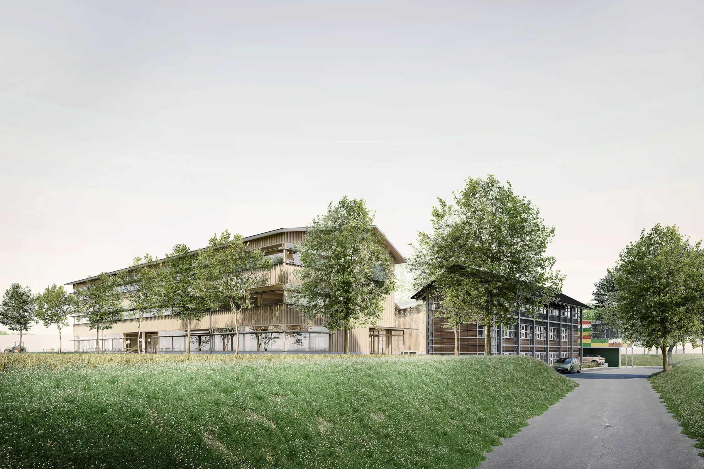 Mehrgeschossiger Holzbau mit vertikaler Lattenfassade steht hinter einer Wiese, davor führt eine Straße vorbei.